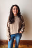 A woman with long dark wavy hair stands indoors wearing a beige knit sweater with cream ribbed trim on the shoulders and cuffs, paired with blue jeans, against a plain off-white wall.
