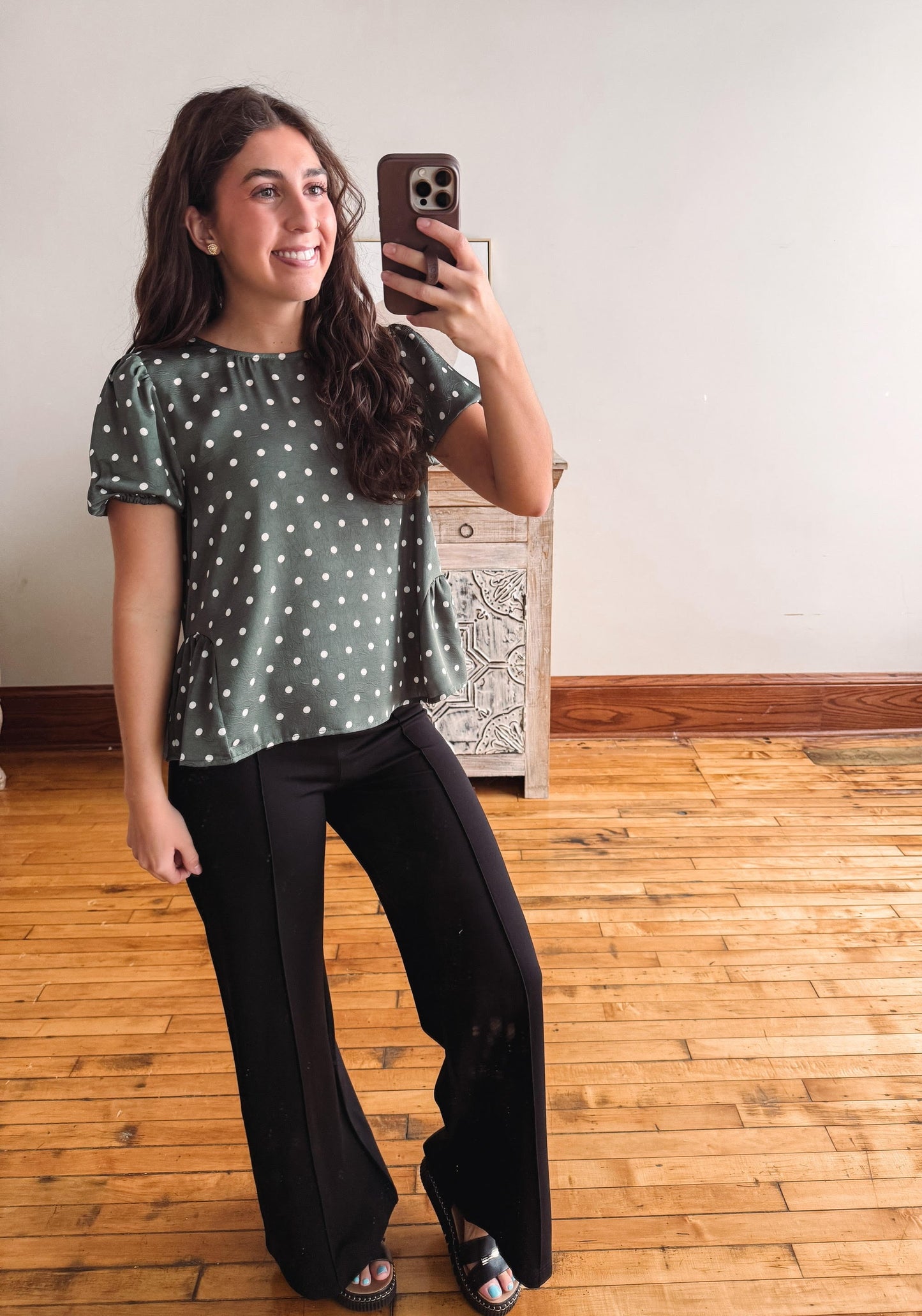 A woman takes a mirror selfie wearing a sage green short-sleeve top with white polka dots and black wide-leg pants in a room with a wooden floor and white wall.