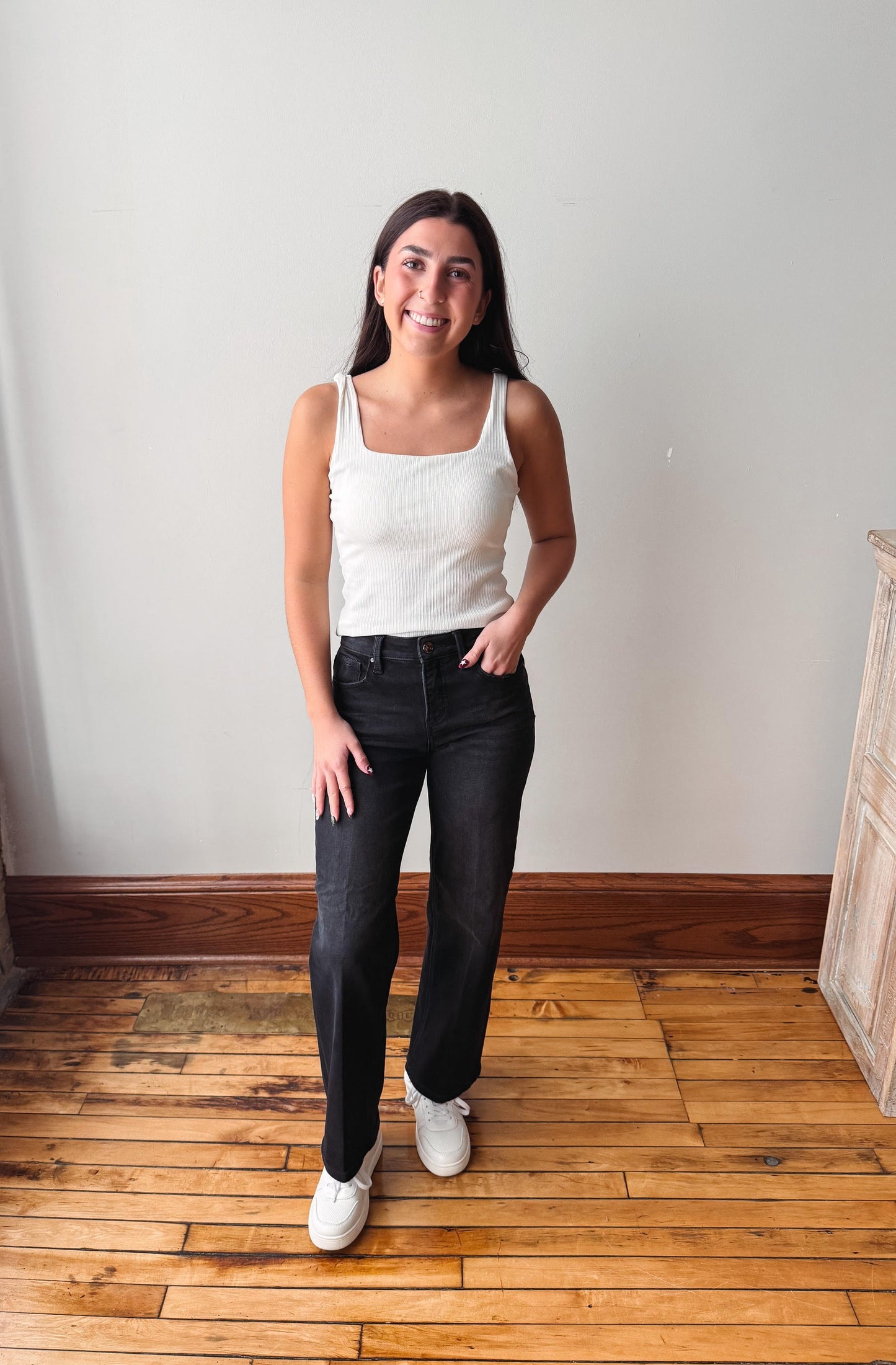 A woman stands in a room with a light gray wall and wooden floor, wearing a white ribbed tank top, black jeans, and white sneakers.