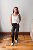 A woman stands in a room with a light gray wall and wooden floor, wearing a white ribbed tank top, black jeans, and white sneakers.