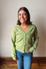 A smiling woman with brown hair stands indoors wearing a light sage green knit cardigan with buttons over a white top and blue jeans, against a plain light wall with a wooden baseboard and floor.