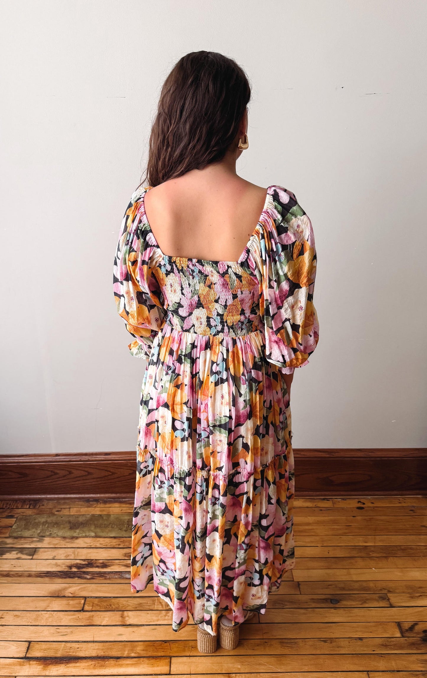 Woman wearing a colorful floral dress standing on a wooden floor with a white wall background