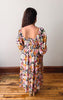 Woman wearing a colorful floral dress standing on a wooden floor with a white wall background