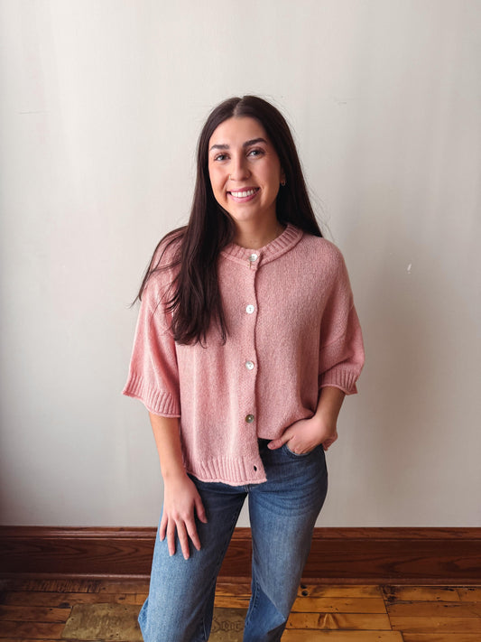 A woman with long dark hair stands indoors wearing a dusty pink short-sleeve button-front cardigan and blue jeans against a plain off-white wall with a wooden baseboard and floor.