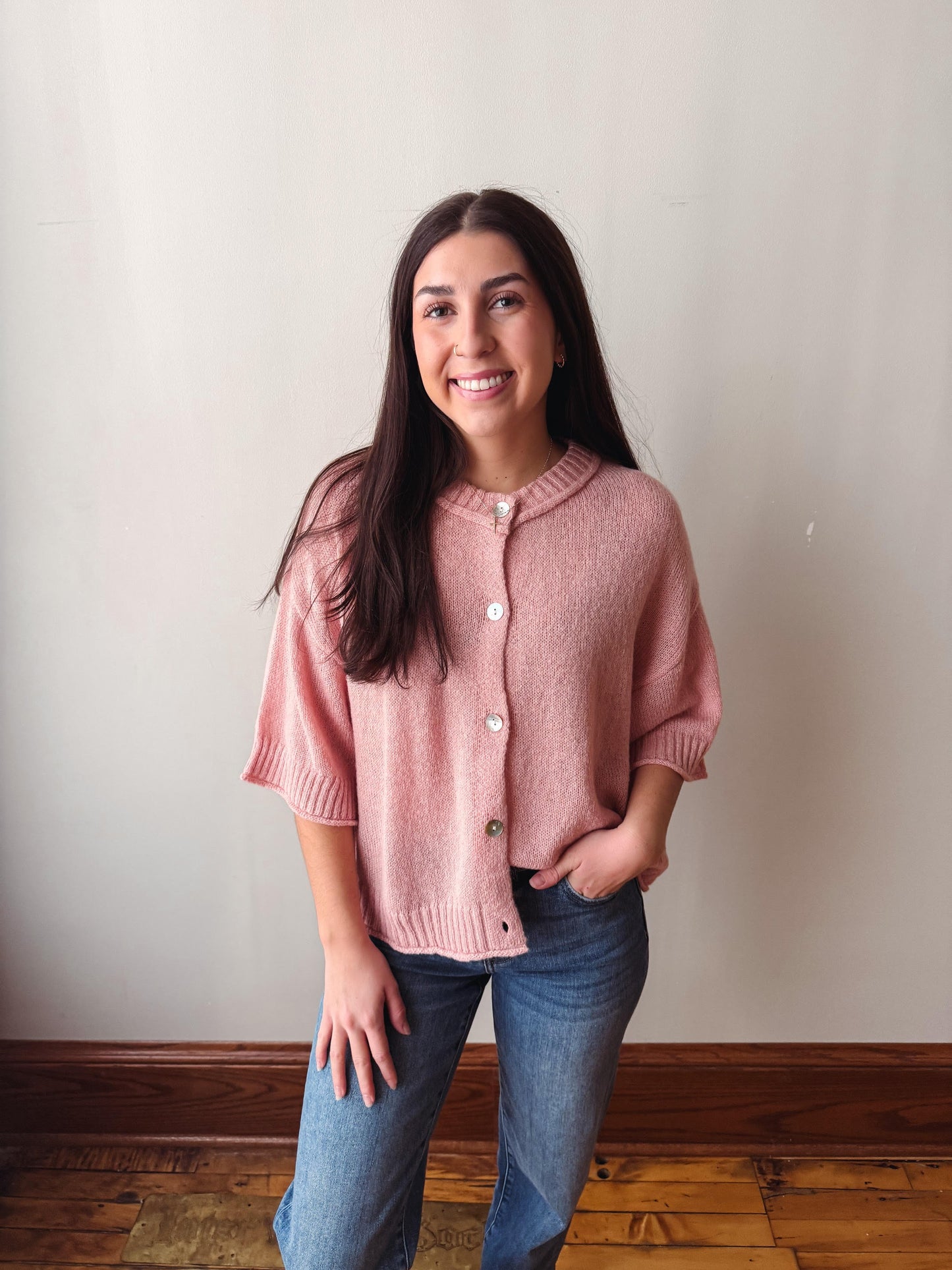 A woman with long dark hair stands indoors wearing a dusty pink short-sleeve button-front cardigan and blue jeans against a plain off-white wall with a wooden baseboard and floor.
