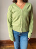 A person wearing a light green knit cardigan with cream buttons and blue jeans, standing indoors against a plain wall and wooden floor.