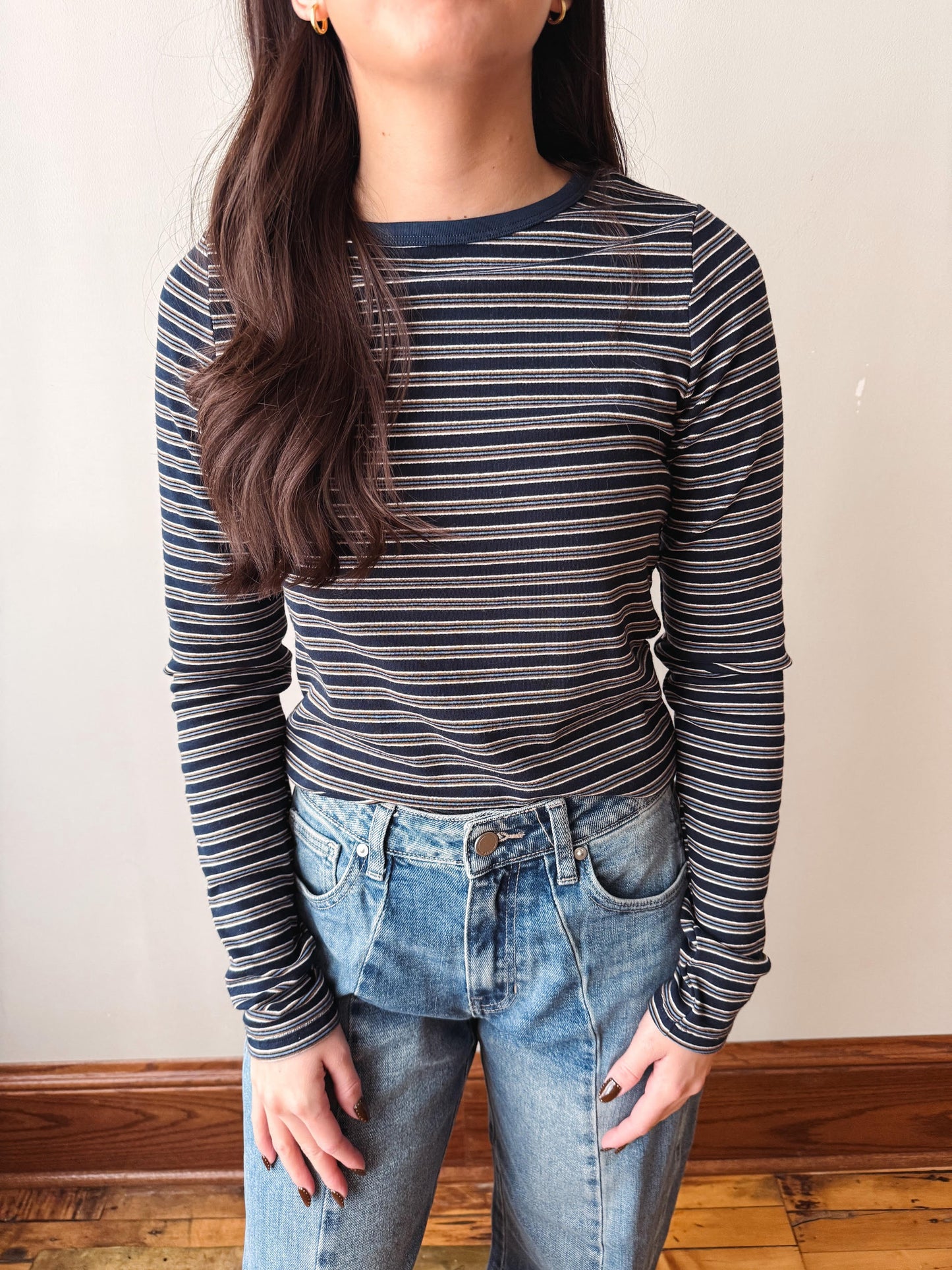 A woman wearing a navy blue and white striped long-sleeve t-shirt with blue jeans, standing against a plain beige wall.