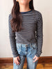 A woman wearing a navy blue and white striped long-sleeve t-shirt with blue jeans, standing against a plain beige wall.