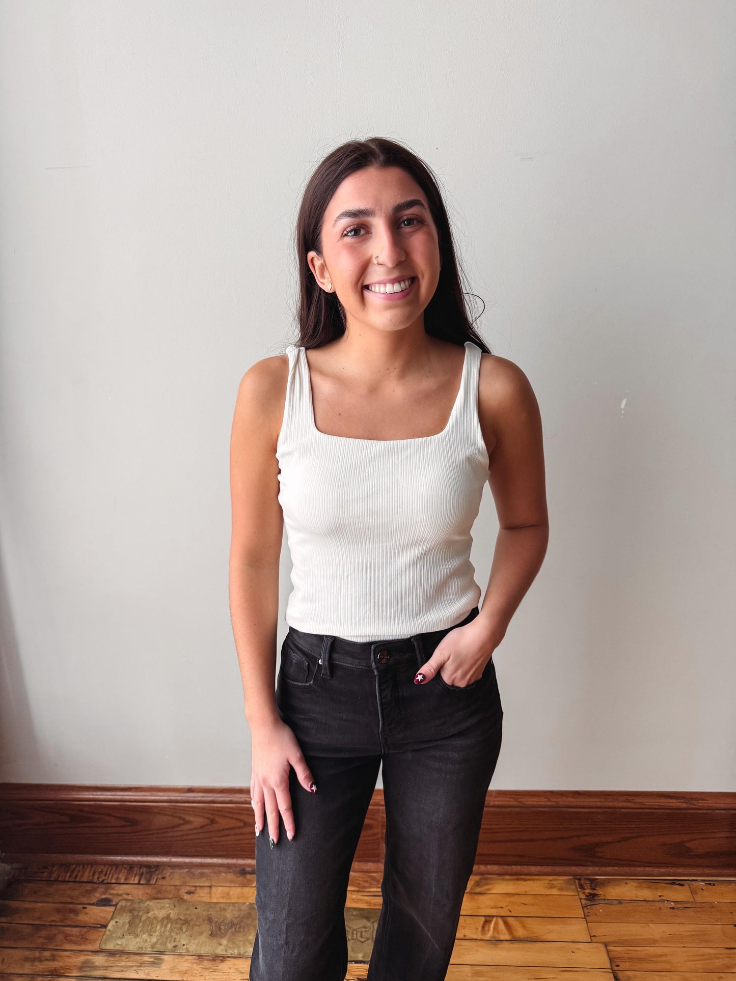 A smiling young woman stands indoors against a plain off-white wall, wearing a fitted white ribbed tank top and dark charcoal jeans.