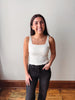 A smiling young woman stands indoors against a plain off-white wall, wearing a fitted white ribbed tank top and dark charcoal jeans.