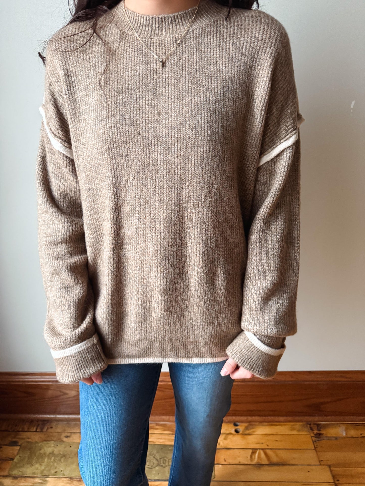 A person wearing a ribbed knit crewneck sweater in taupe-beige with contrasting cream trim on the sleeve cuffs and shoulder yokes, paired with blue jeans.