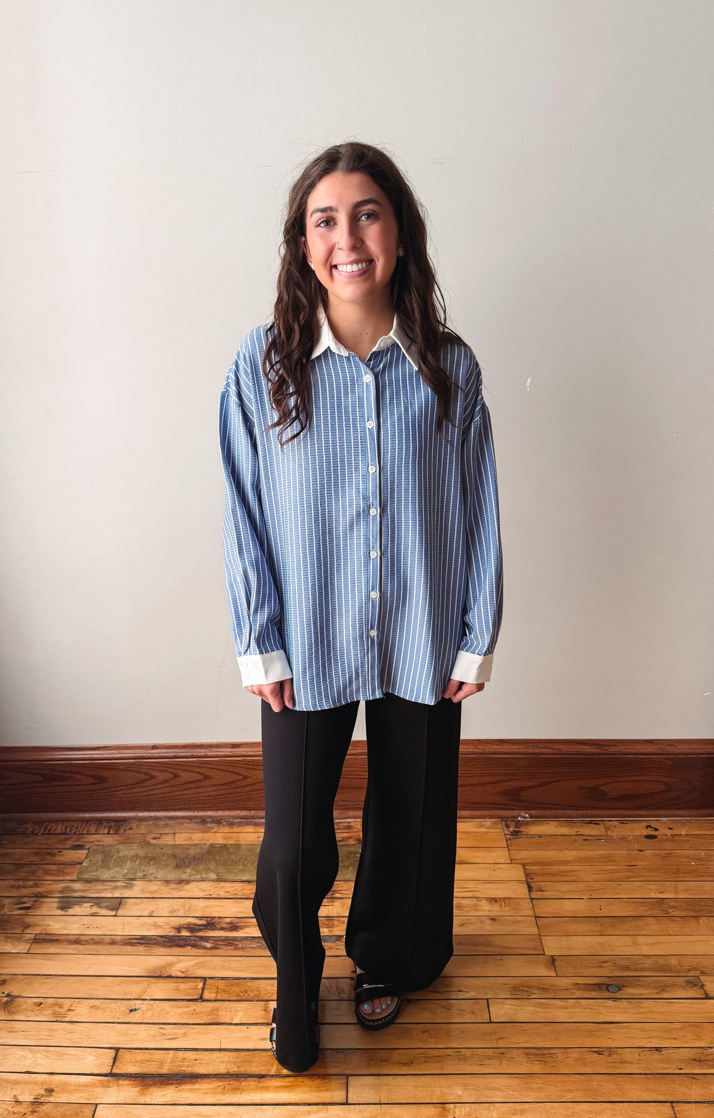 A woman stands in a room with a beige wall and wooden floor, wearing a blue and white vertically striped button-down shirt with black wide-leg pants.