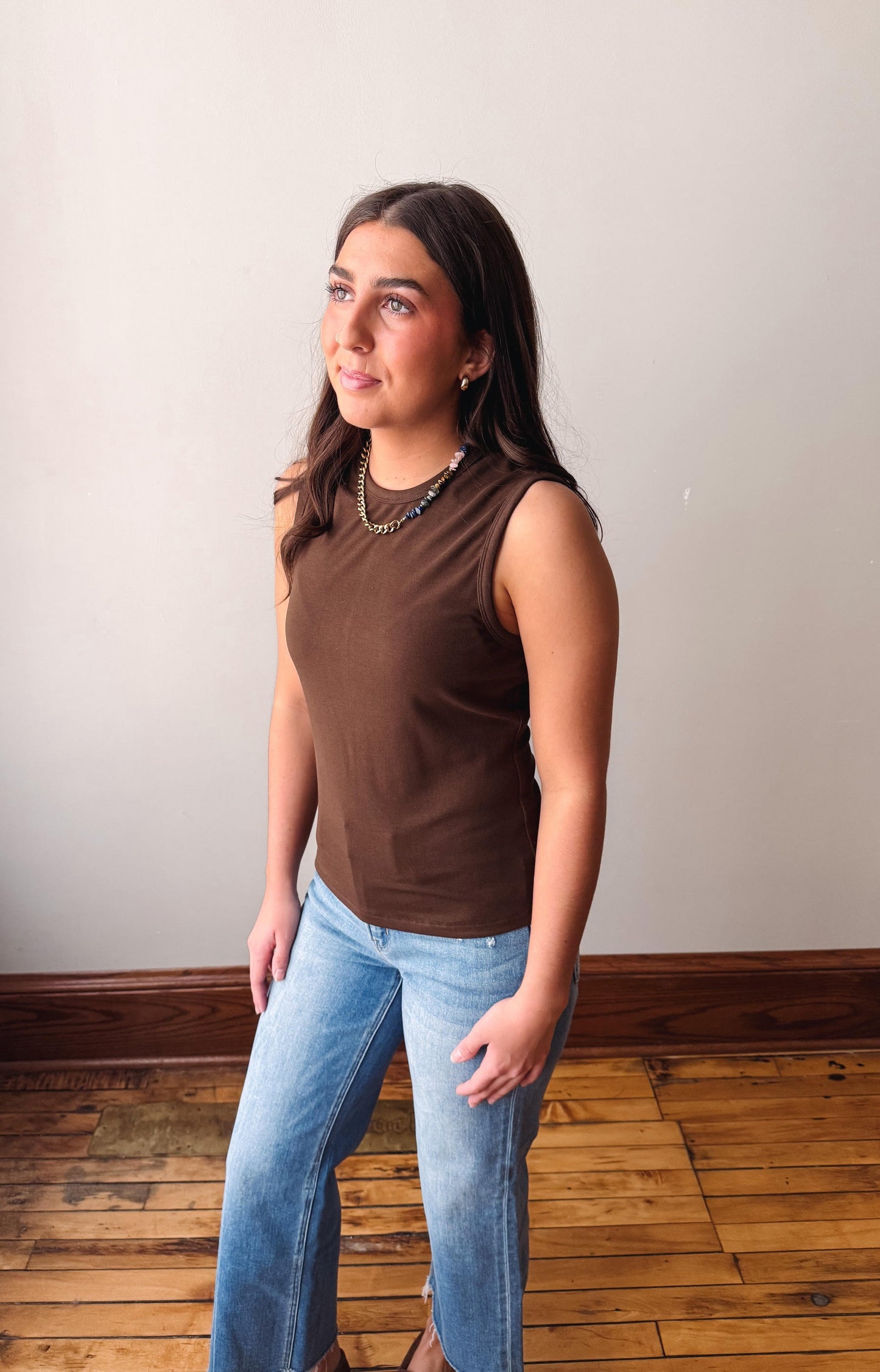 Woman wearing a brown sleeveless top and blue jeans standing against a plain wall.