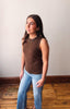 Woman wearing a brown sleeveless top and blue jeans standing against a plain wall.