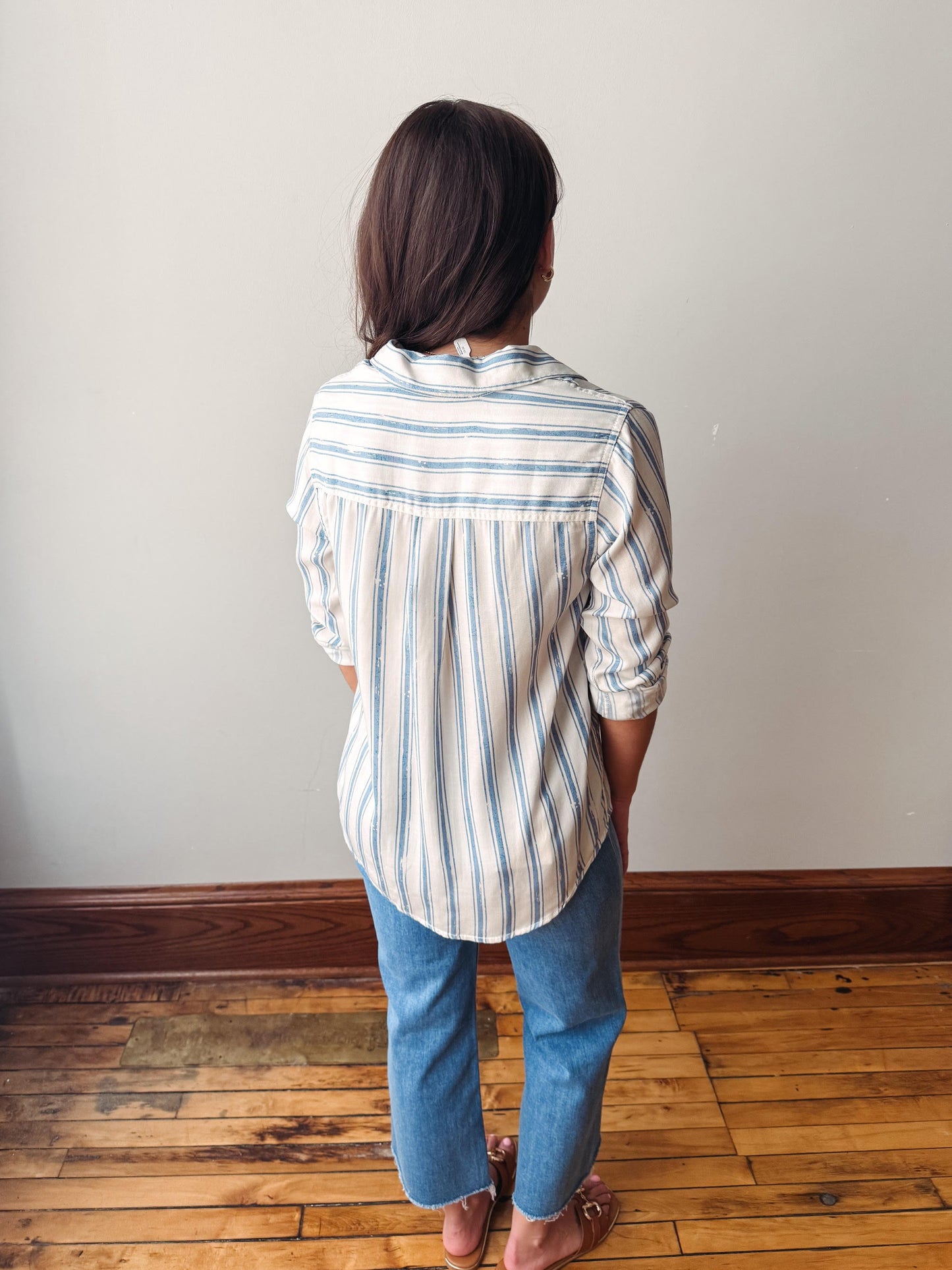 Person wearing a striped shirt and jeans standing on a wooden floor against a white wall.