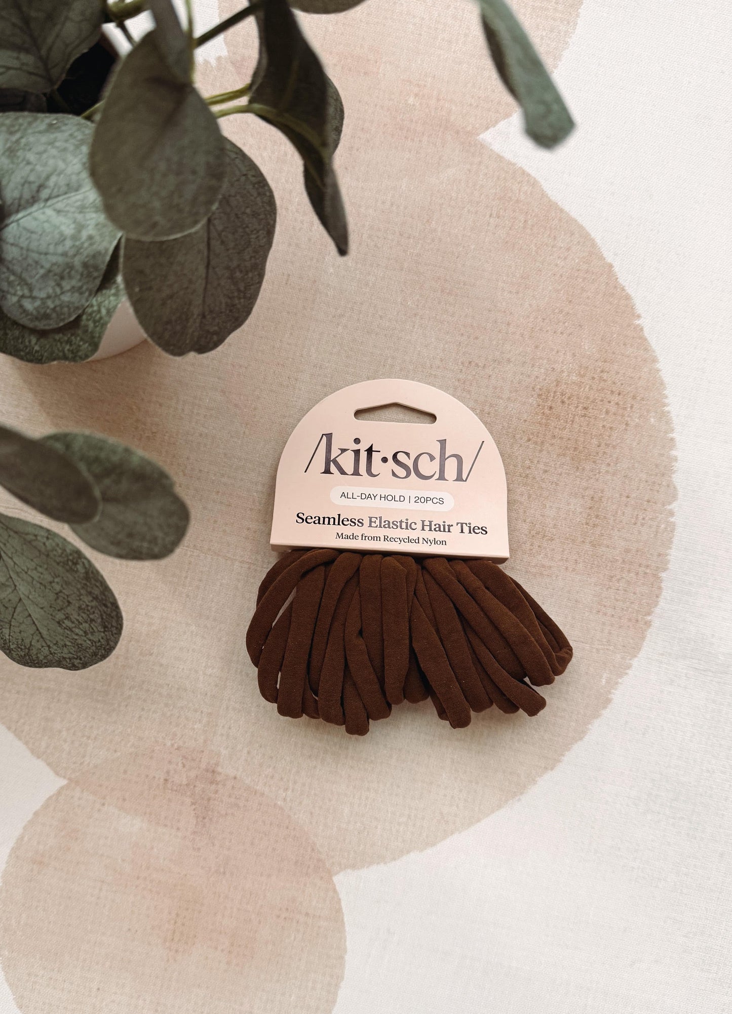 A pack of Kit-sch seamless elastic hair ties in a dark brown color, attached to a light pink cardboard hanger. The product is styled on a textured beige background with watercolor circles and green eucalyptus leaves.