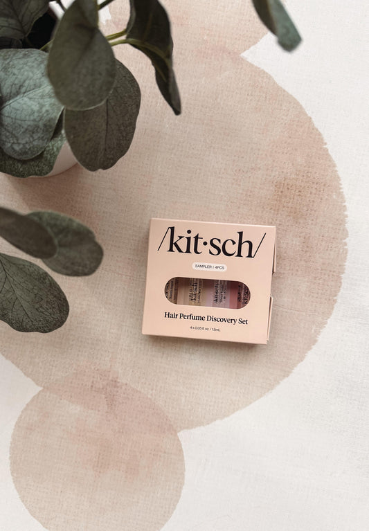A Kit Sch Hair Perfume Discovery Set box with a clear window showing three pink hair perfume tubes inside, styled on a textured beige surface with a plant leaf in the corner.