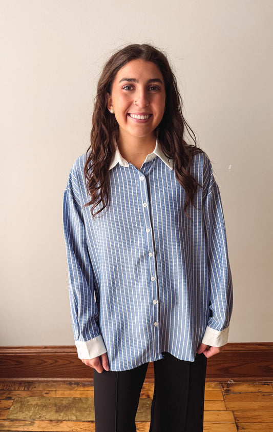 A woman with long dark hair stands indoors wearing a light blue and white vertically striped button-down shirt with a white collar and cuffs, paired with black pants.