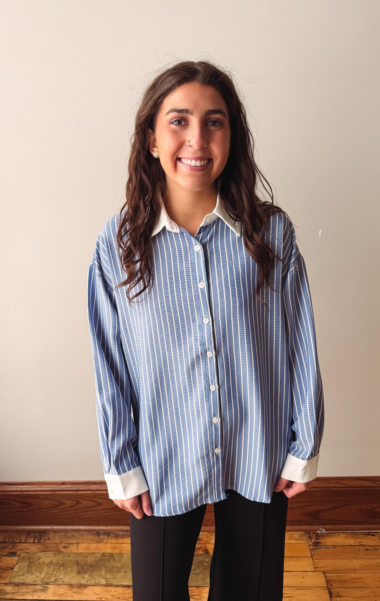 A woman with long dark hair stands indoors wearing a light blue and white vertically striped button-down shirt with a white collar and cuffs, paired with black pants.