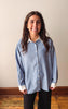 A woman with long dark hair stands indoors wearing a light blue and white vertically striped button-down shirt with a white collar and cuffs, paired with black pants.