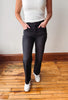 A person wearing dark charcoal gray straight-leg jeans, a white ribbed tank top, and white sneakers, standing on a wooden floor against a plain wall.