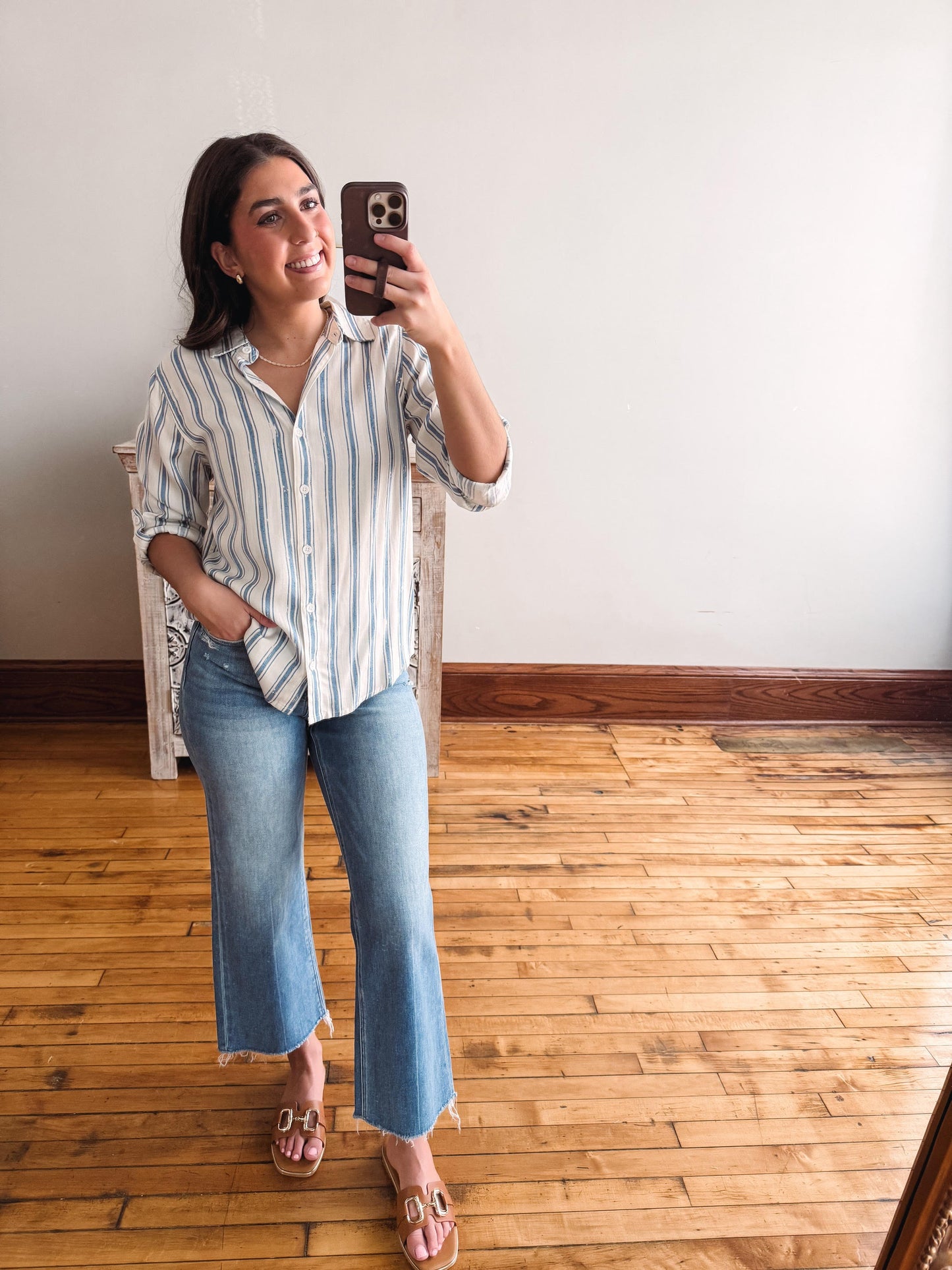 Woman taking a mirror selfie wearing a striped shirt and jeans in a room with wooden flooring.