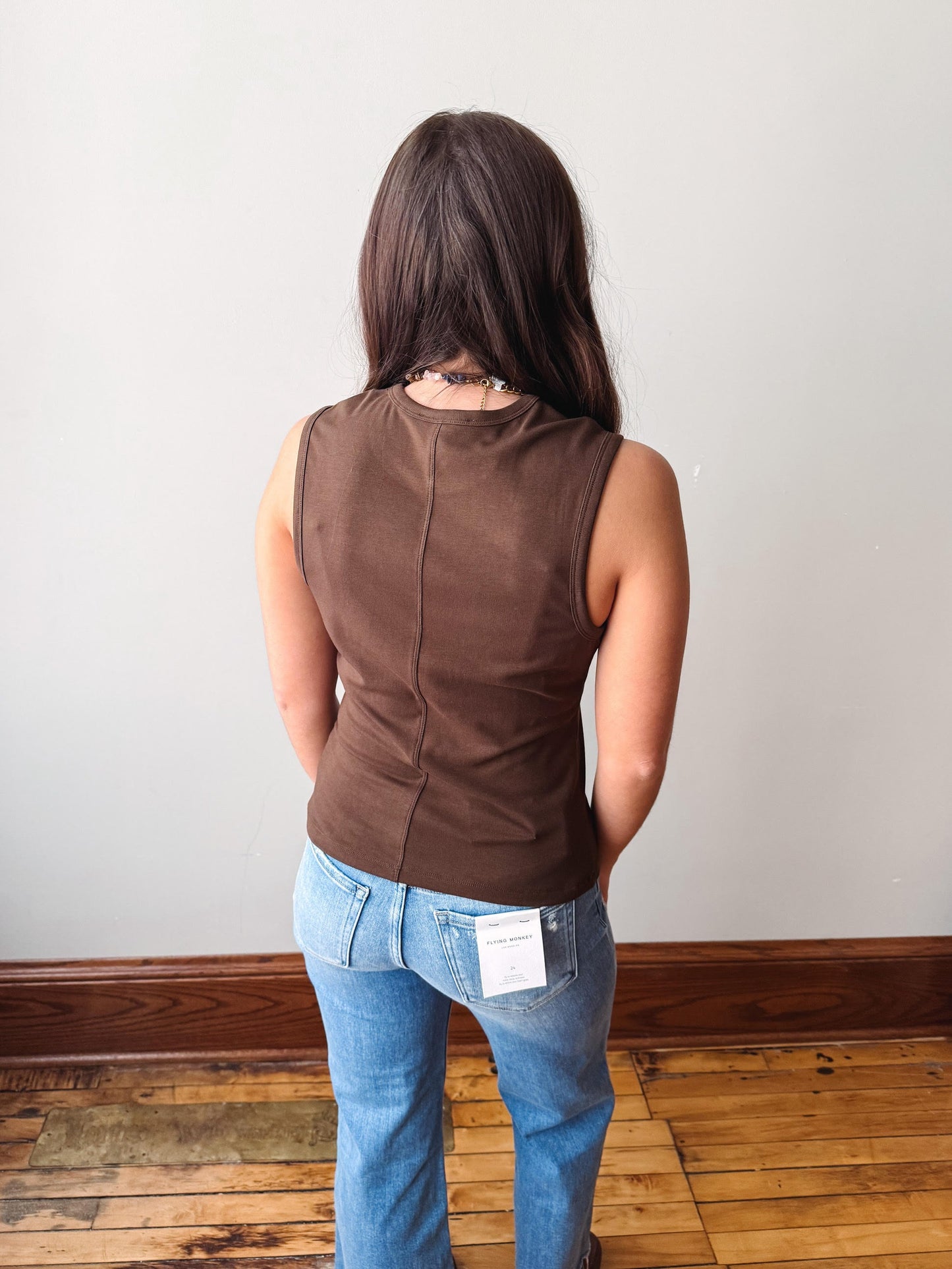 Person wearing a brown sleeveless top and blue jeans standing against a plain wall.
