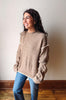 A woman wearing a beige knit sweater with white contrast trim and blue jeans, standing in a room with a light-colored wall and wooden floor.