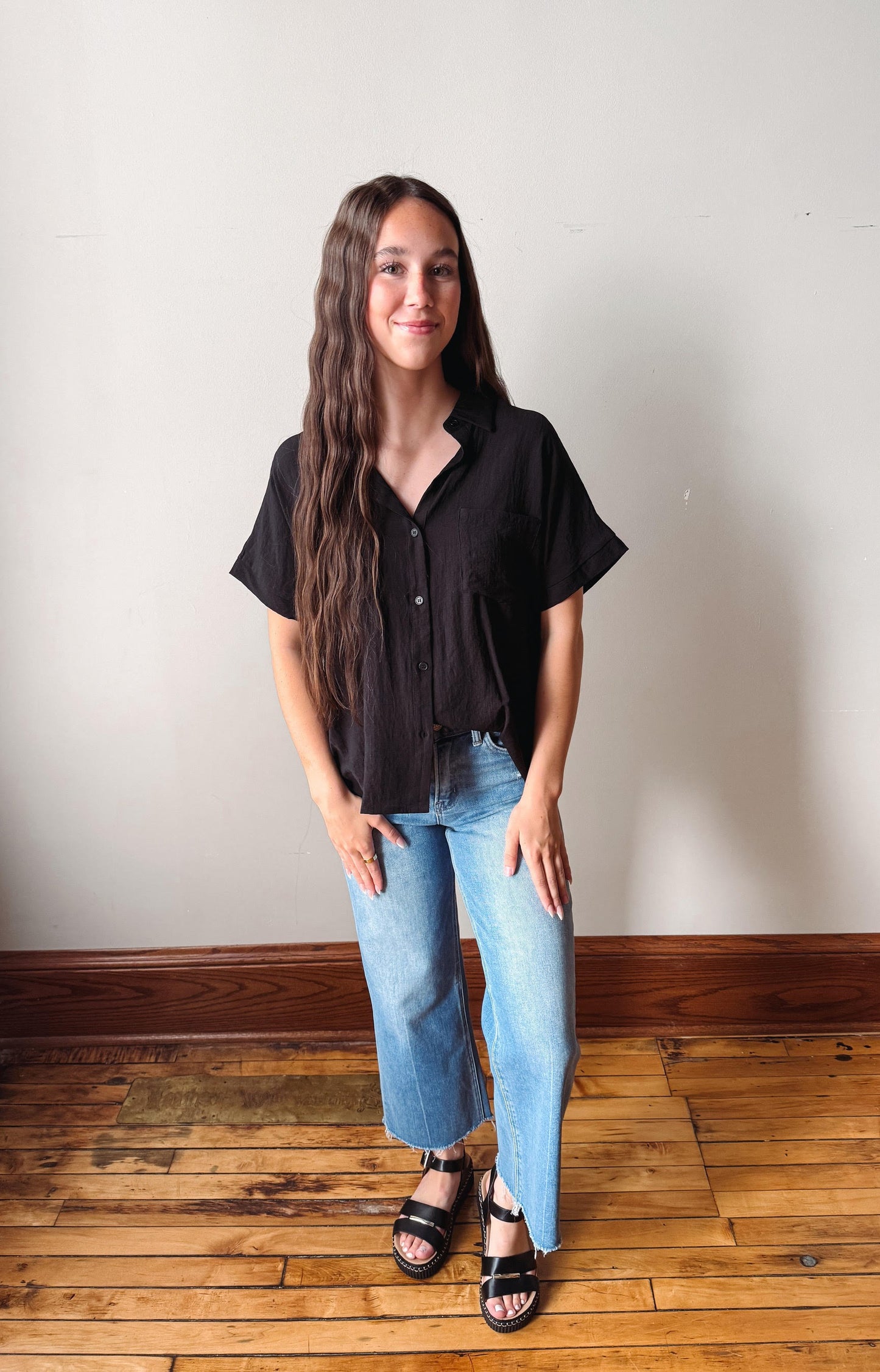 Woman wearing a black shirt and blue jeans standing on a wooden floor with a white wall background