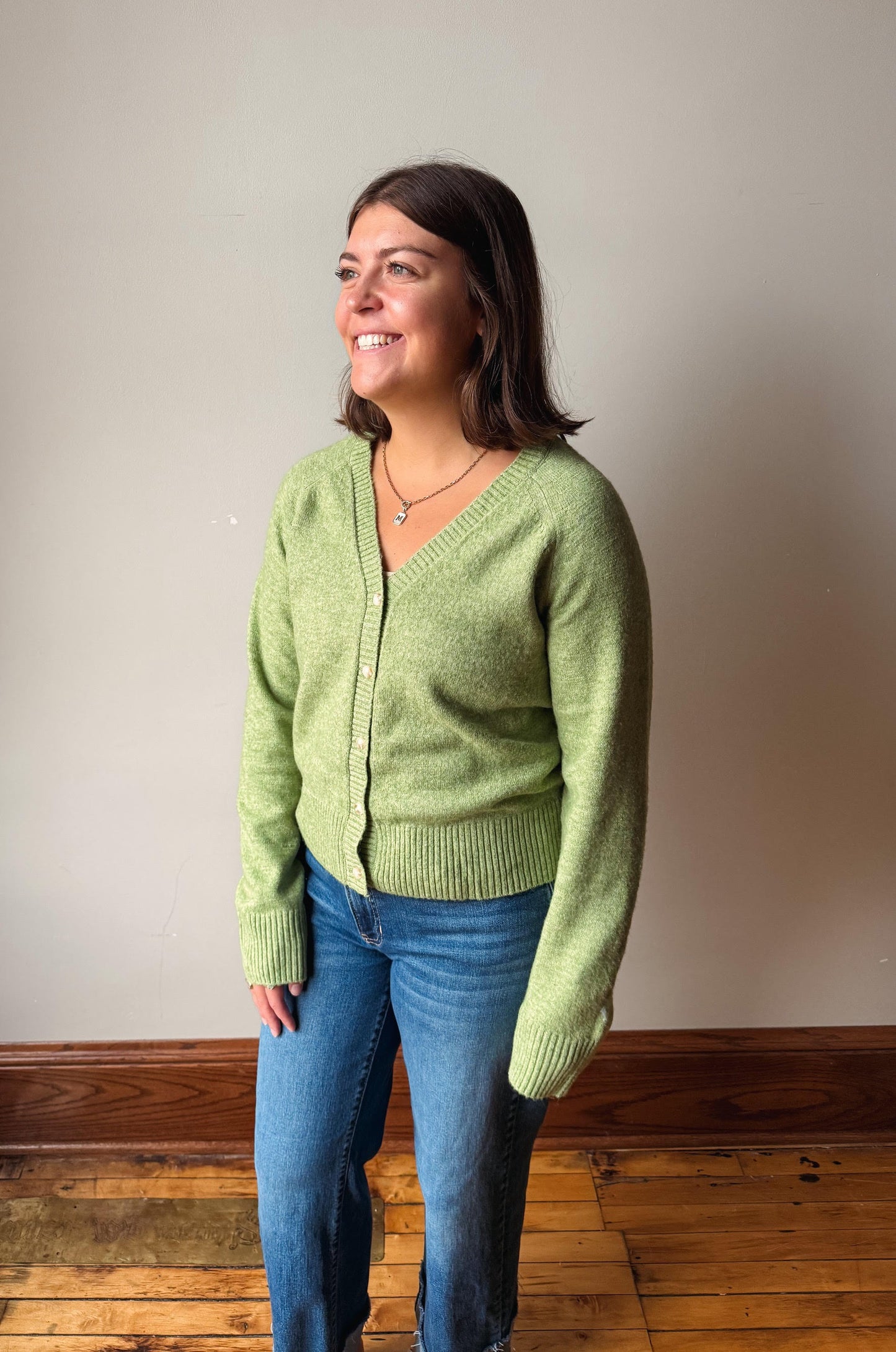 A woman wearing a light green knit cardigan and blue jeans, standing in a room with a beige wall and wooden floor.