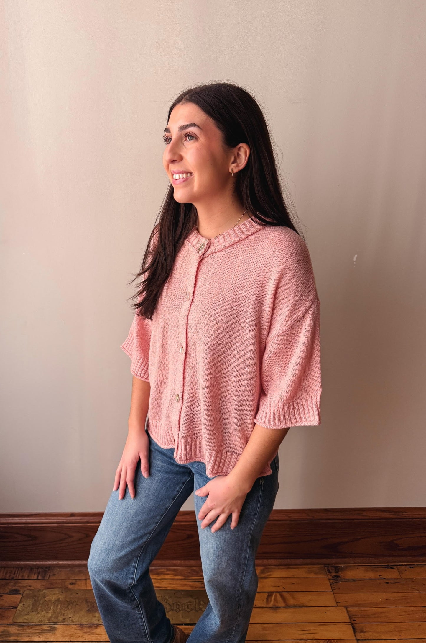 A woman wearing a light pink knit cardigan with elbow-length sleeves and blue jeans, standing in a room with a beige wall and wooden floor.