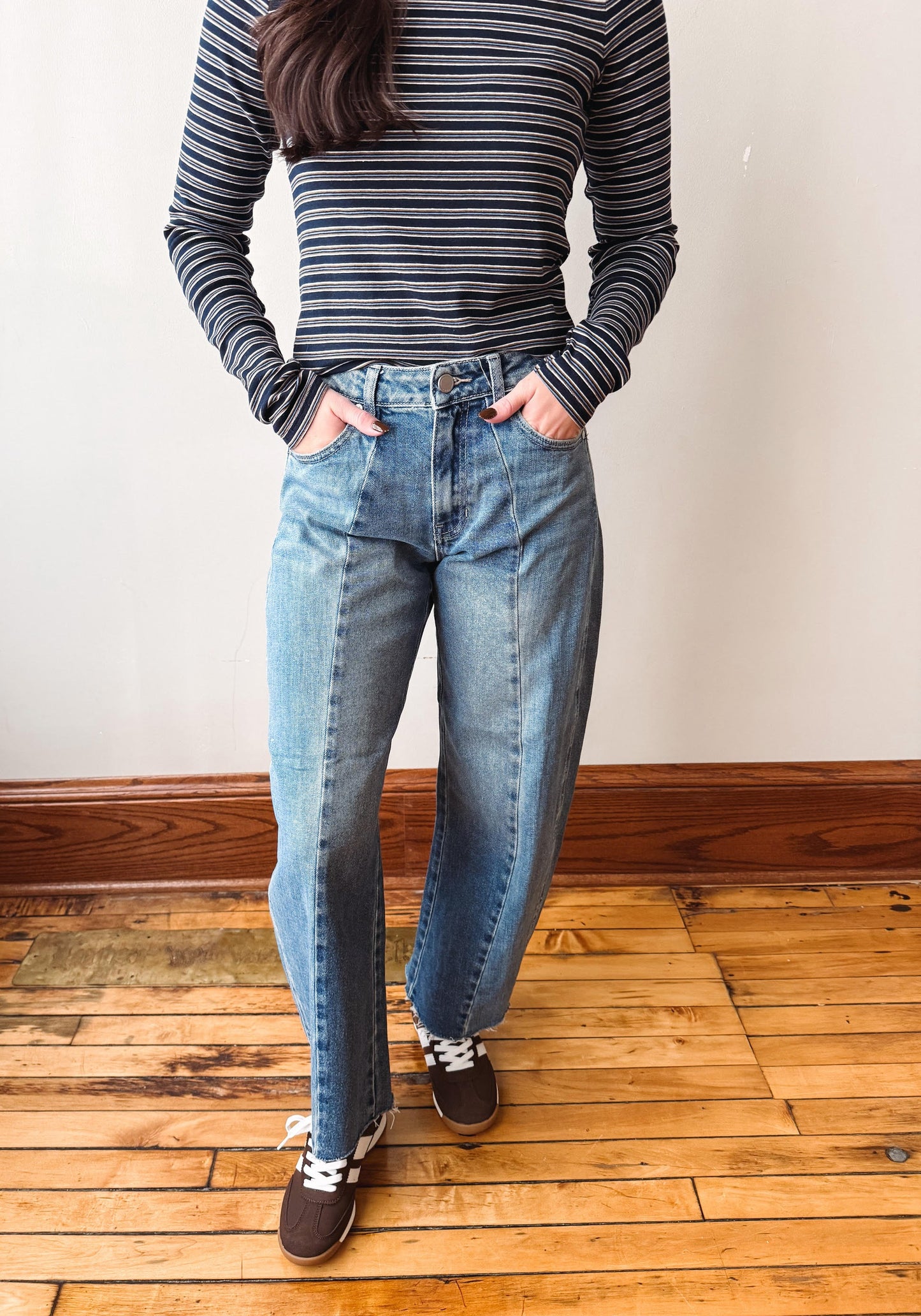A person wearing a navy and gray striped long-sleeve shirt and light-wash blue jeans, standing on a wooden floor against a plain wall.