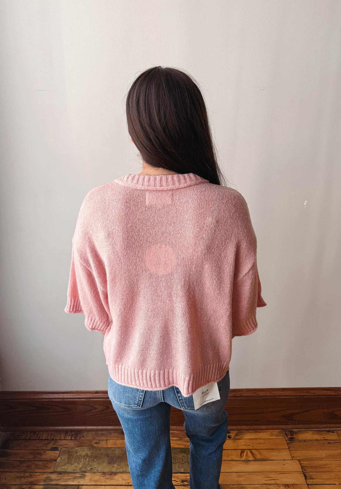 A woman viewed from behind wearing a light pink knit sweater and blue jeans, standing on a wooden floor against a plain off-white wall.