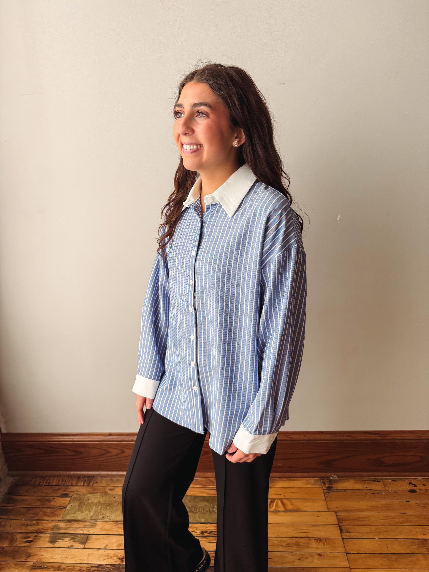 A woman wearing a blue and white vertically striped button-down shirt with a white collar and cuffs, paired with black pants, standing in a room with a beige wall and wooden floor.