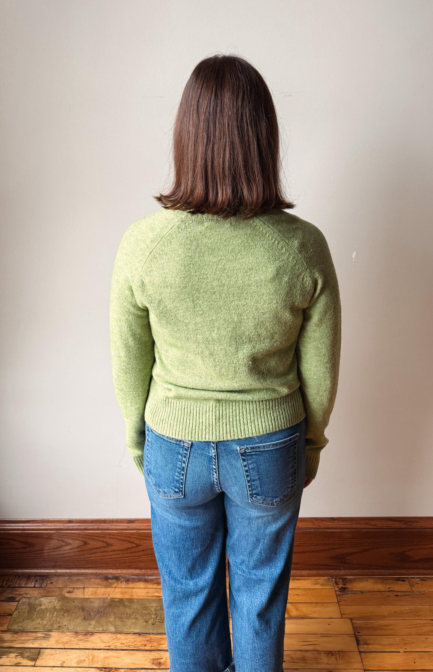 A woman viewed from behind wearing a light green knit sweater and blue jeans, standing on a wooden floor against a plain off-white wall.