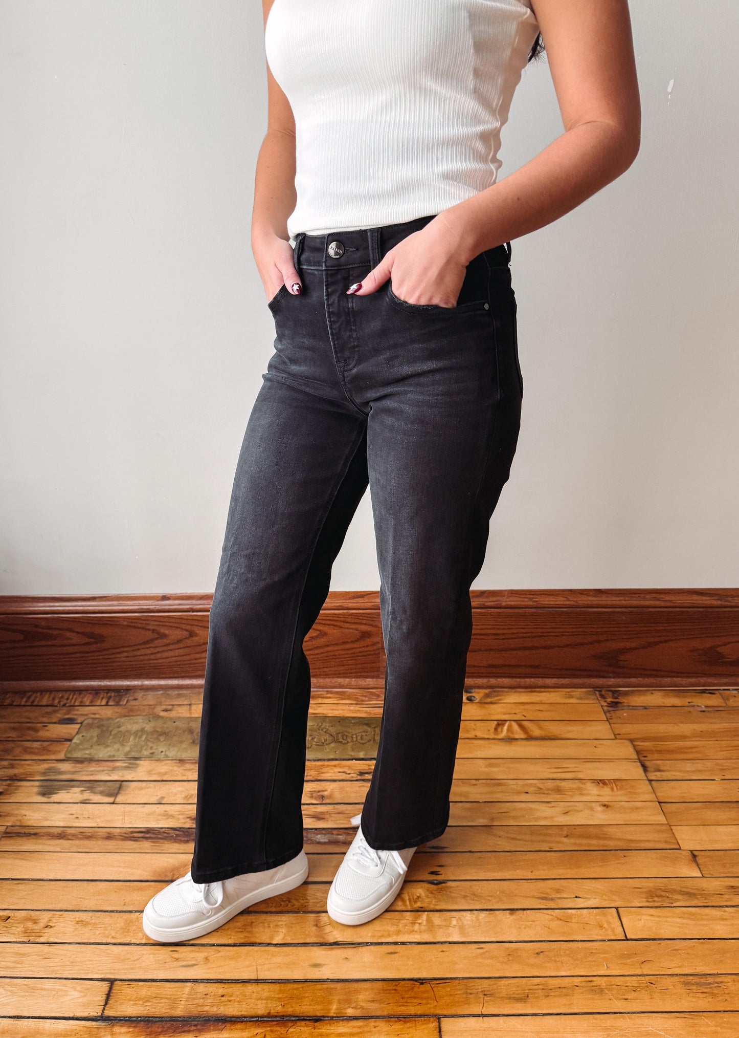 A person wearing dark charcoal gray wide-leg jeans, a white ribbed tank top, and white sneakers, standing on a wooden floor against a plain wall.