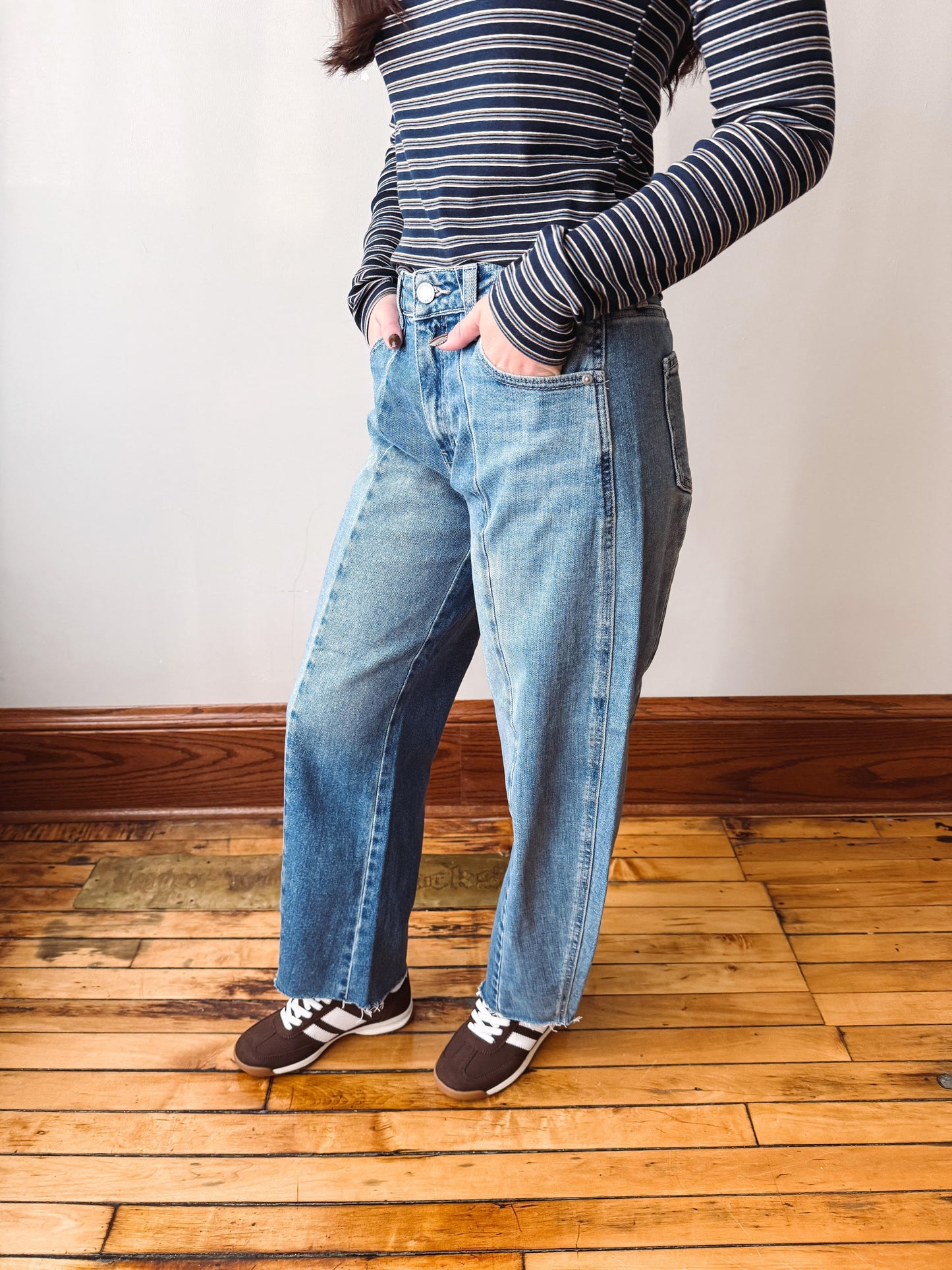 A person wearing light-wash blue straight-leg jeans, a navy and white striped long-sleeve top, and brown sneakers, standing on a wooden floor against a plain wall.