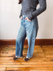 A person wearing light-wash blue straight-leg jeans, a navy and white striped long-sleeve top, and brown sneakers, standing on a wooden floor against a plain wall.