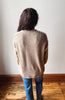 A person viewed from behind wearing a beige knit sweater with contrasting cream shoulder panels and blue jeans, standing on a wooden floor against a plain off-white wall.