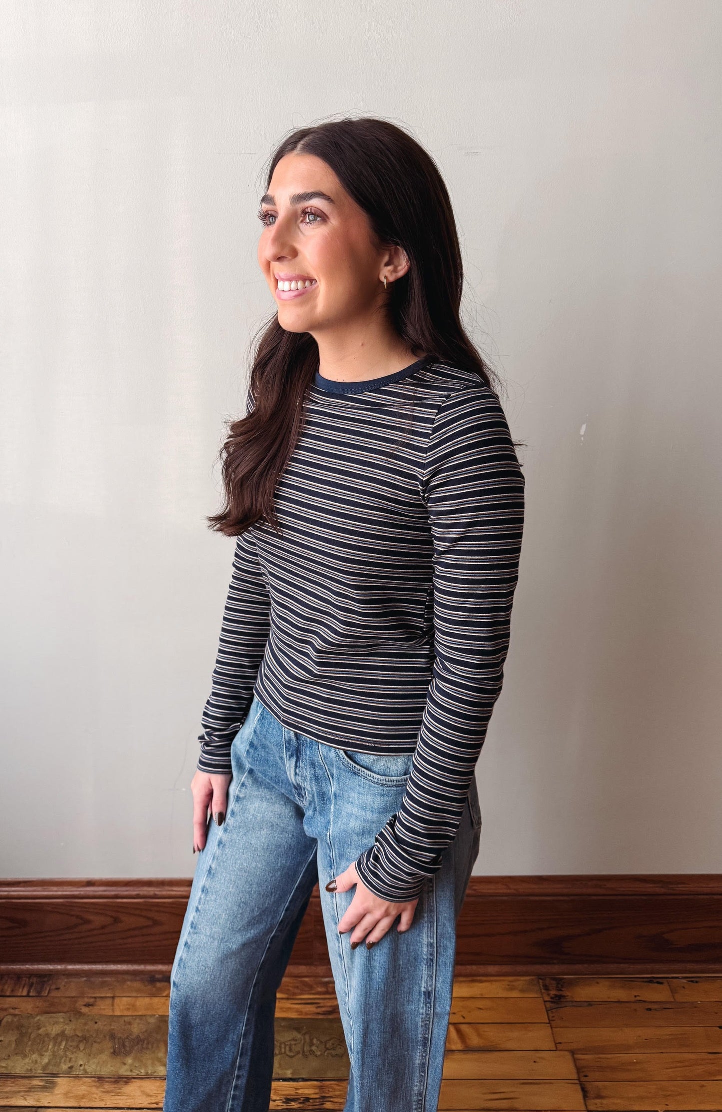 A woman with long dark hair is shown in profile wearing a long-sleeve crewneck shirt with thin horizontal navy and gray stripes, paired with light-wash blue jeans, standing against a plain off-white wall with a wooden baseboard and floor.