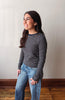 A woman with long dark hair is shown in profile wearing a long-sleeve crewneck shirt with thin horizontal navy and gray stripes, paired with light-wash blue jeans, standing against a plain off-white wall with a wooden baseboard and floor.