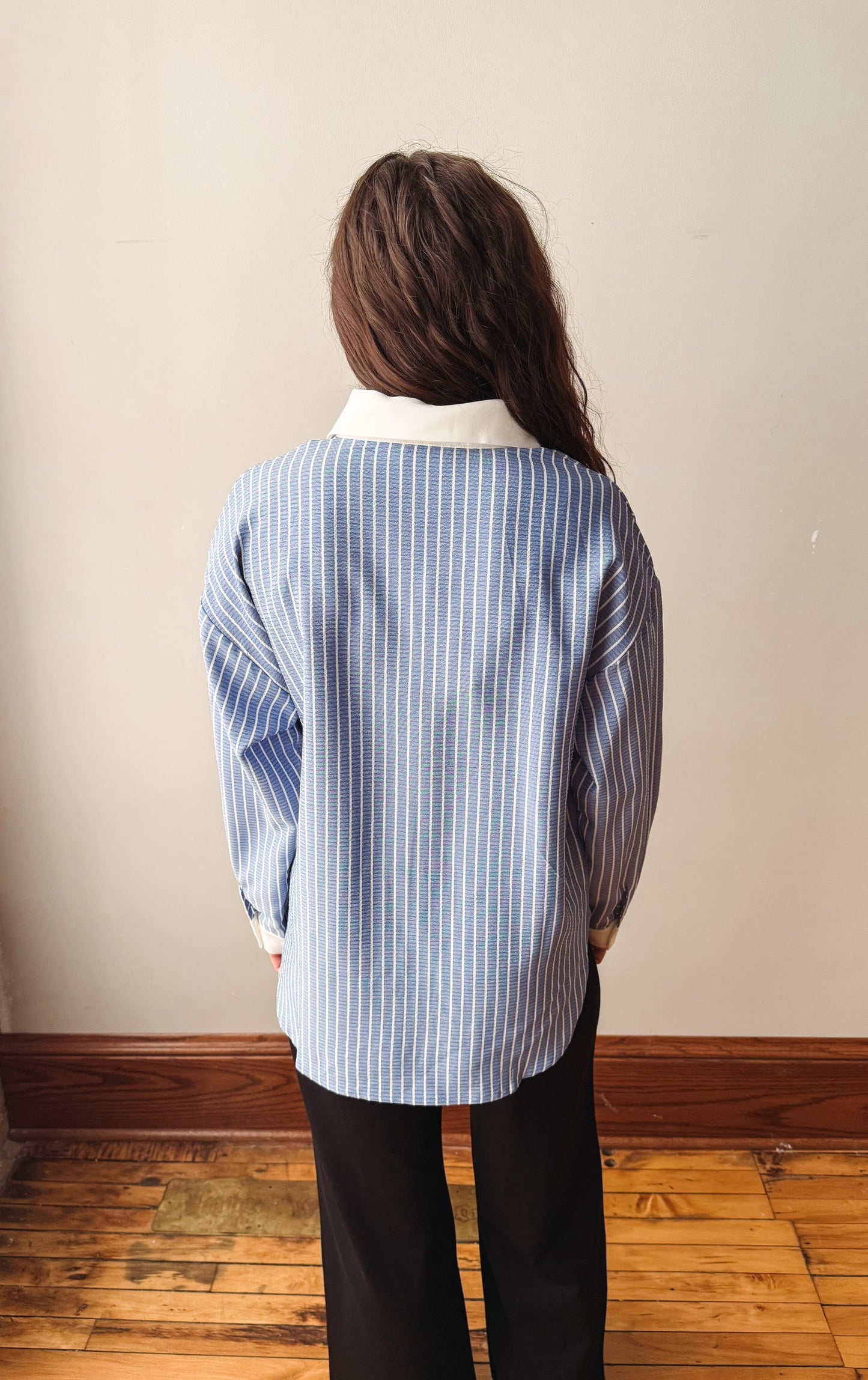 A person viewed from behind wearing a light blue and white vertically striped button-up shirt with a white collar, paired with black pants, standing in an indoor room with a beige wall and wooden floor.