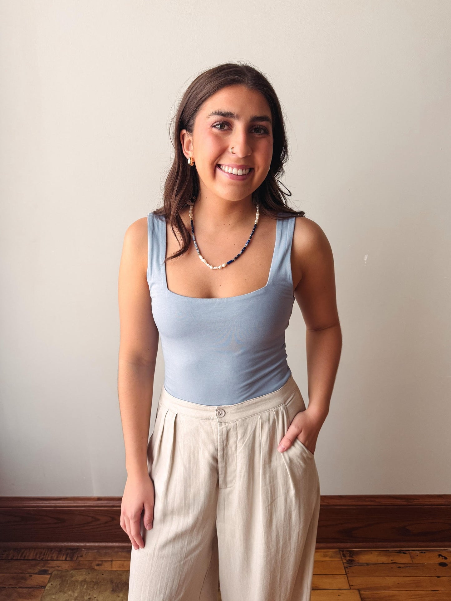 Woman wearing a light blue tank top and beige pants against a plain wall.