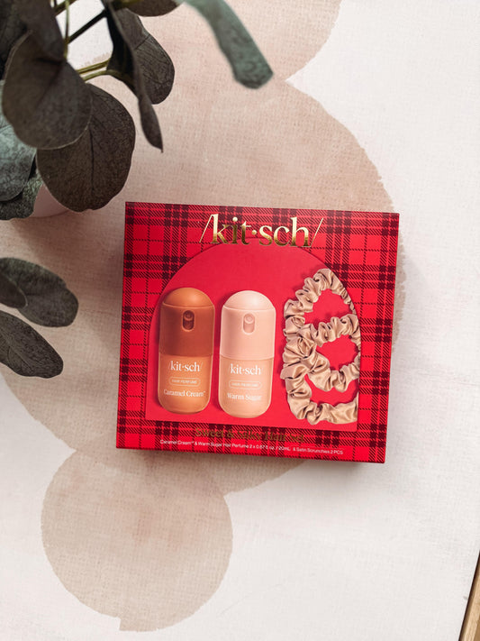 A Kit-sch gift set featuring a red plaid box with two bottles and a scrunchie, styled on a beige surface with decorative leaves.
