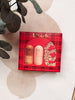 A Kit-sch gift set featuring a red plaid box with two bottles and a scrunchie, styled on a beige surface with decorative leaves.