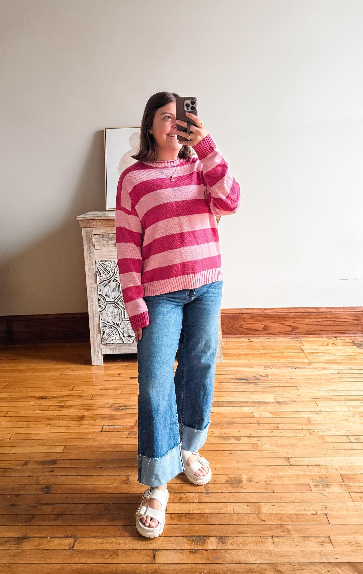 A woman takes a mirror selfie wearing a pink and magenta striped knit sweater, blue cuffed jeans, and white slide sandals in a room with a wood floor and light-colored wall.