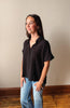 Woman wearing a black shirt and blue jeans standing against a plain wall.