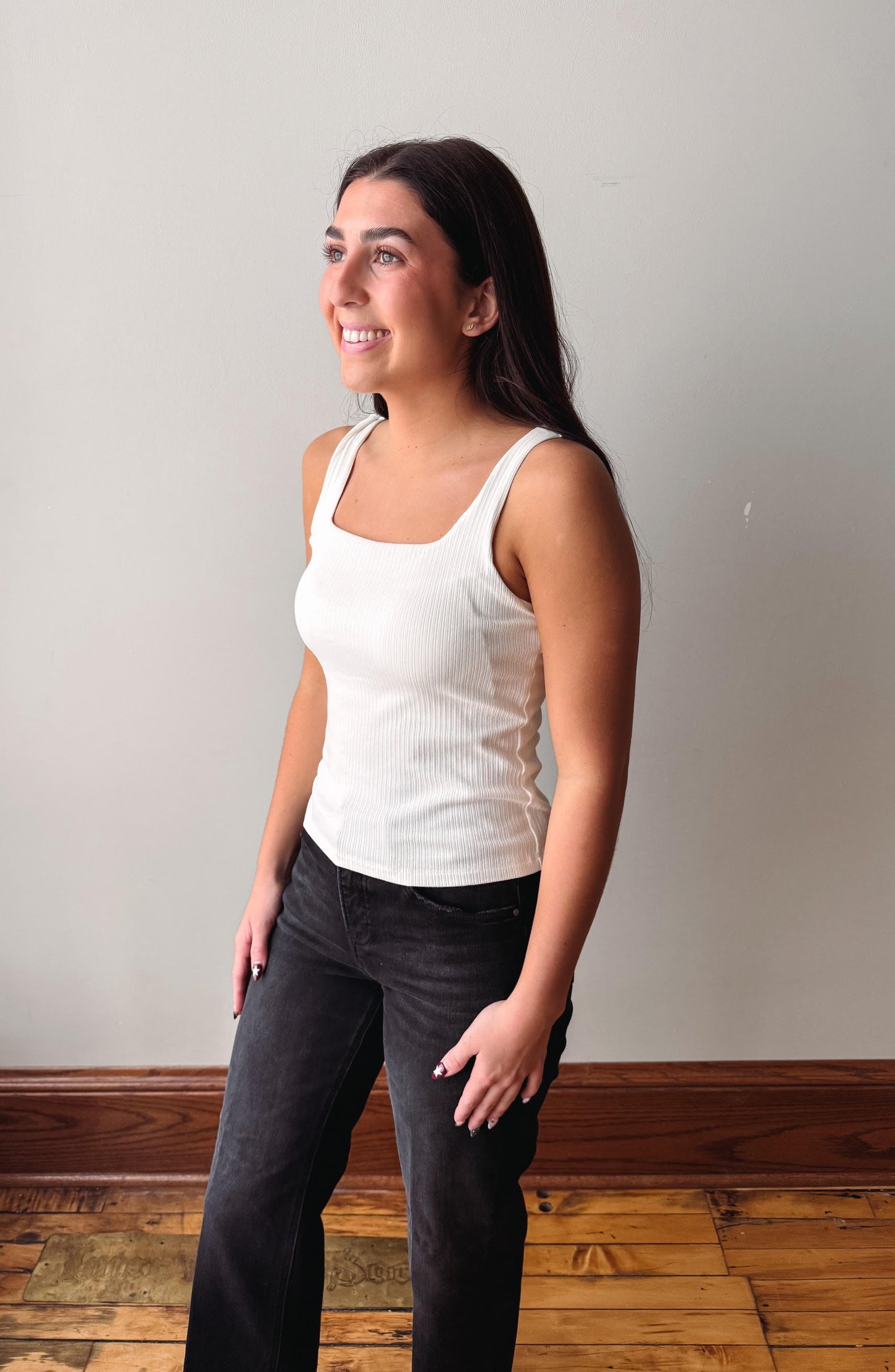 A woman wearing a white ribbed tank top and dark gray jeans, standing in a room with a light gray wall and wooden floor.