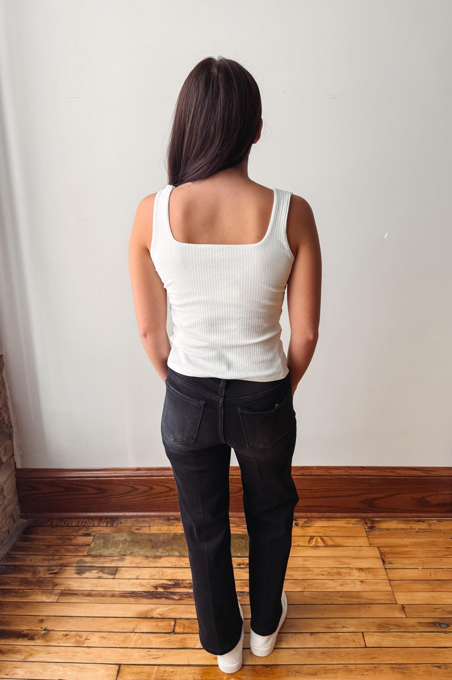 A woman is shown from behind wearing a white ribbed tank top and dark wash jeans, standing on a wooden floor against a plain white wall.