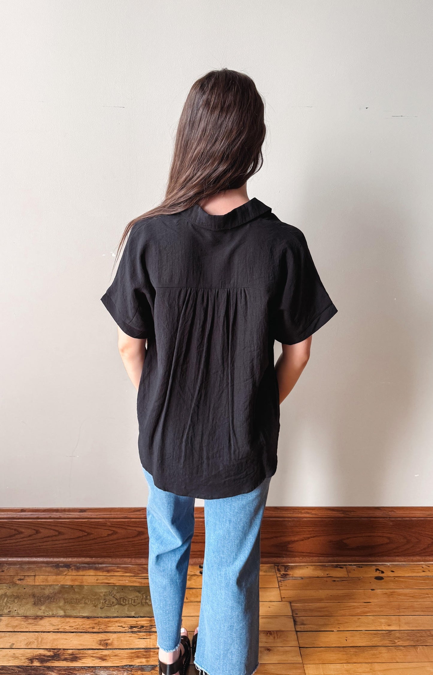 Person wearing a black shirt and blue jeans standing against a white wall on a wooden floor.